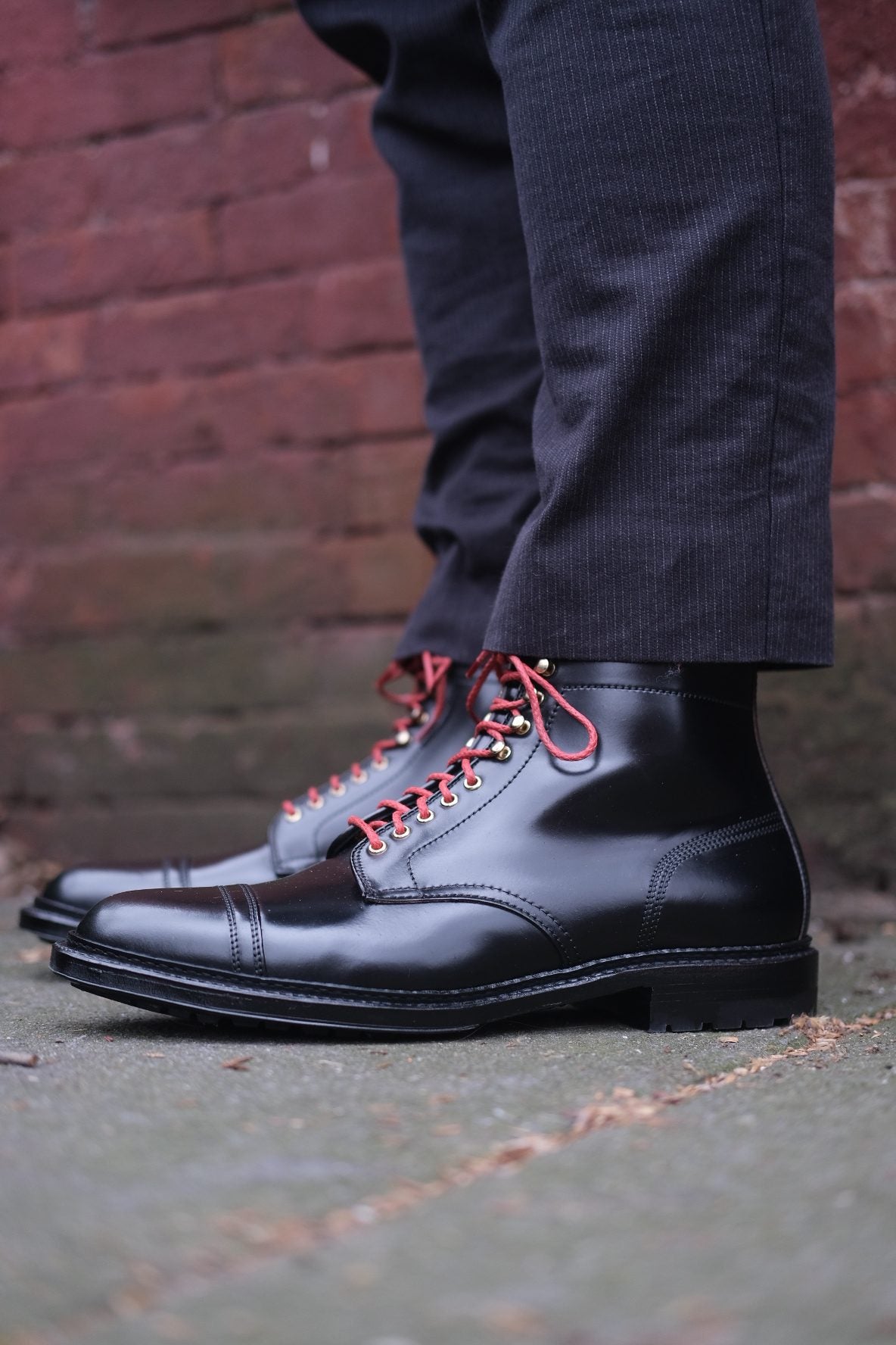 A person stands on stone pavement, sporting Alden Madisons D9867HC Stitchup 2/2 Boots in Black Shell Cordovan with red laces and a commando sole. Theyre dressed in dark pants, exuding a Stitchdown.com Style vibe, against a brick wall background.