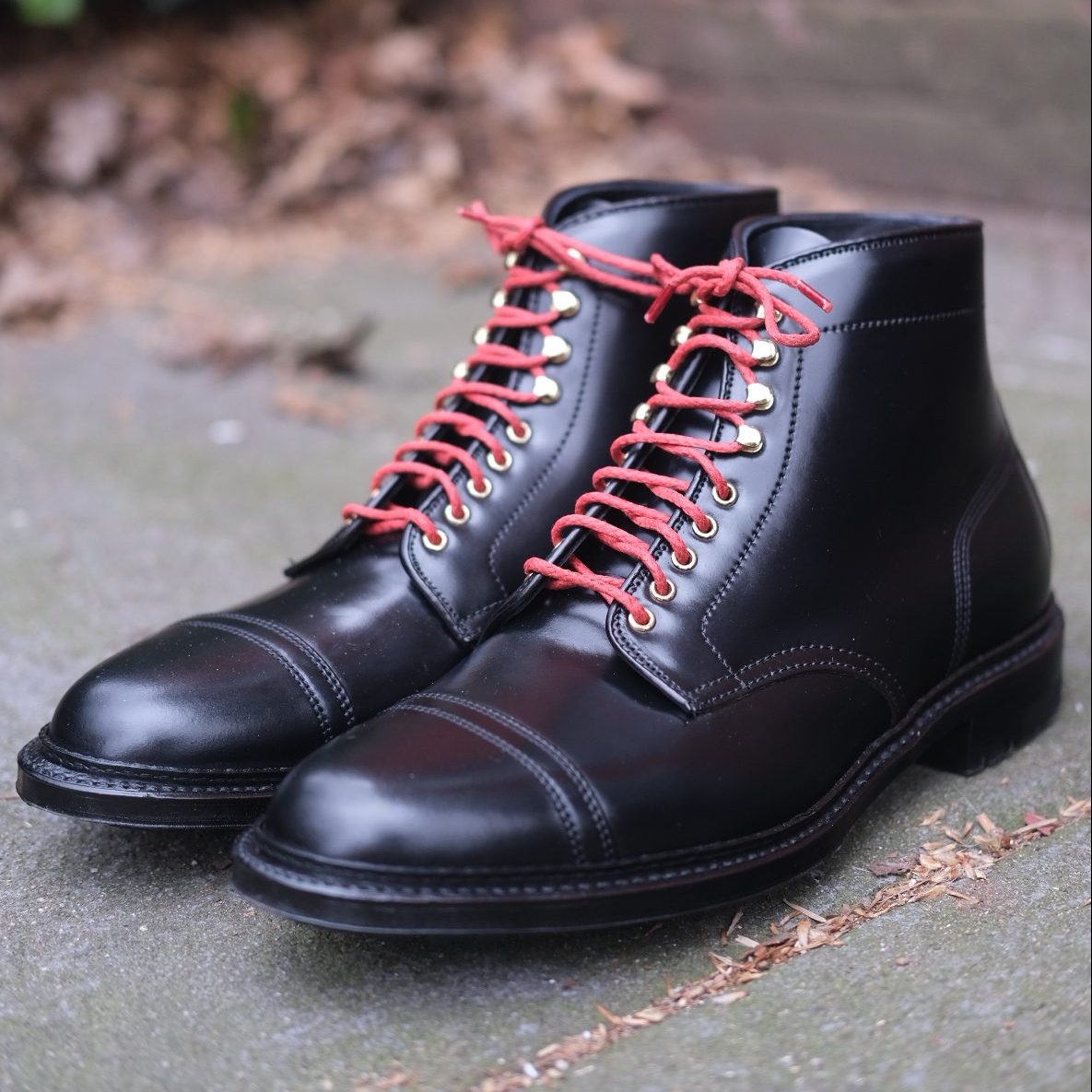 The D9867HC-Stitchup 2/2 Boots by Alden Madison, crafted from Black Shell Cordovan, feature polished black leather with striking red laces and gold eyelets. They rest on a stone surface amidst dry leaves, boasting a rugged Commando Sole for ultimate durability through any adventure.