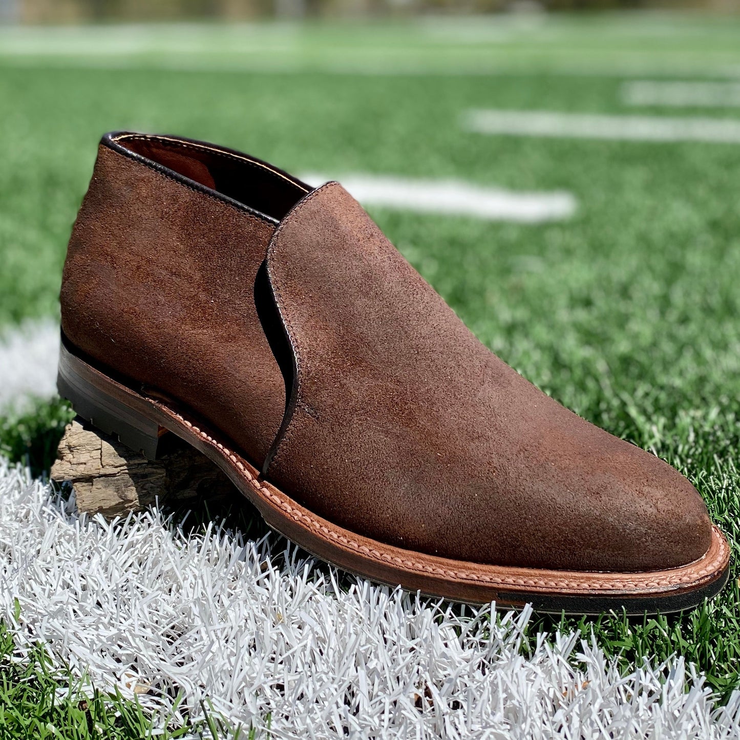 The Alden Madison D8704C - Folsom Slip On Chukka in Tobacco Reverse Chamois is shown on a football field, resting on wood with crisp white yard lines in the background.