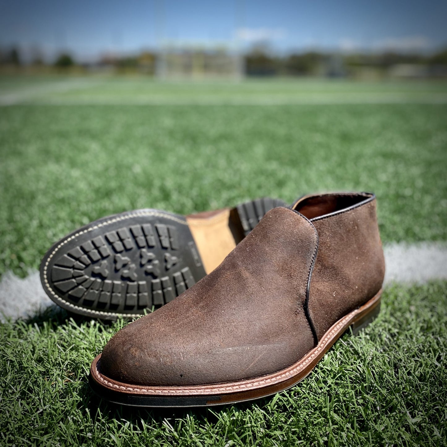 The Alden Madison D8704C Folsom Slip On Chukka in Tobacco Reverse Chamois is shown with its Commando rubber soles, one boot upright and one on its side, on green turf against a blurred sports field backdrop.