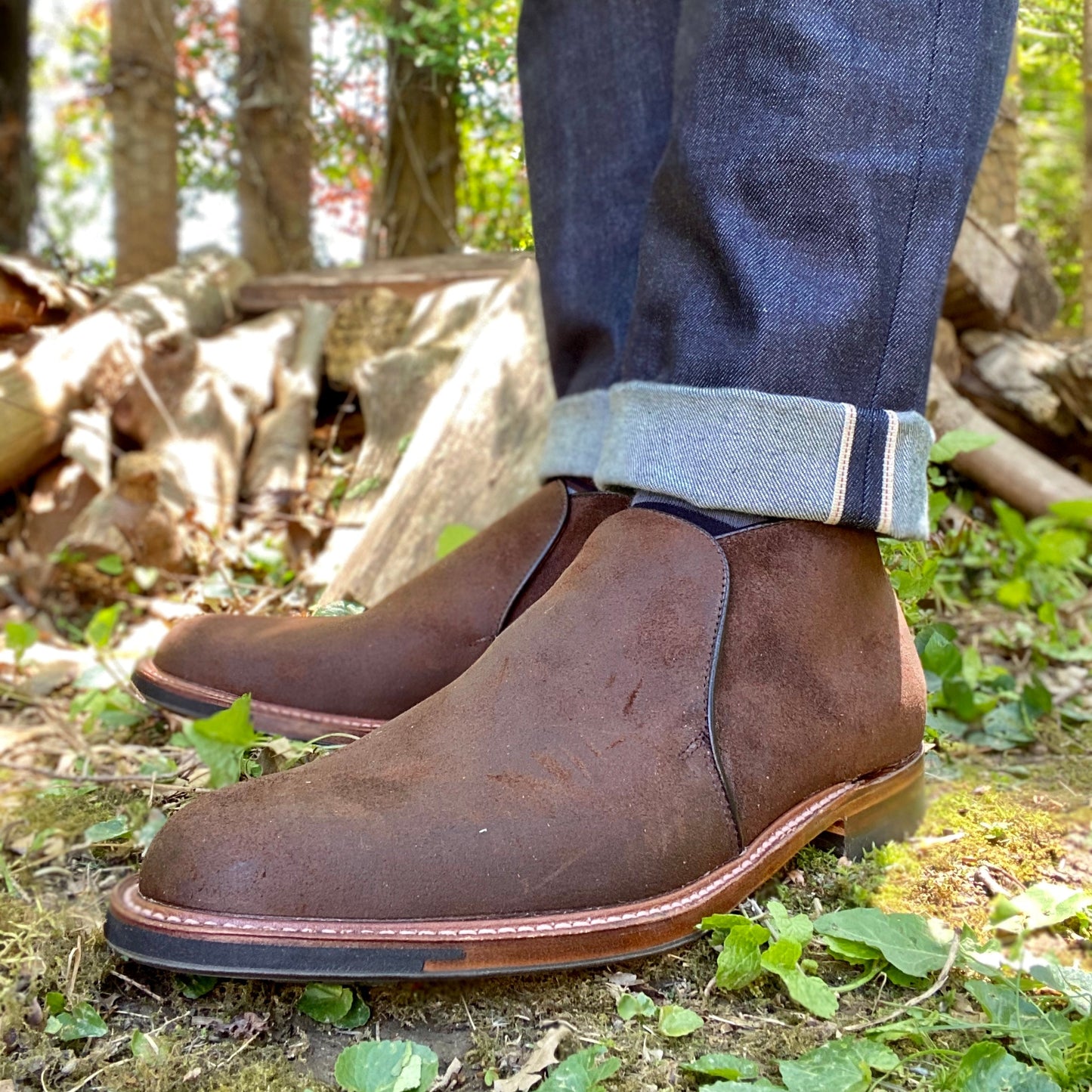 Close-up of a person wearing Alden Madison D8704C - Folsom Slip On Chukka in Tobacco Reverse Chamois with cuffed dark blue jeans, standing on grass and leaves outdoors with logs and trees in the background.