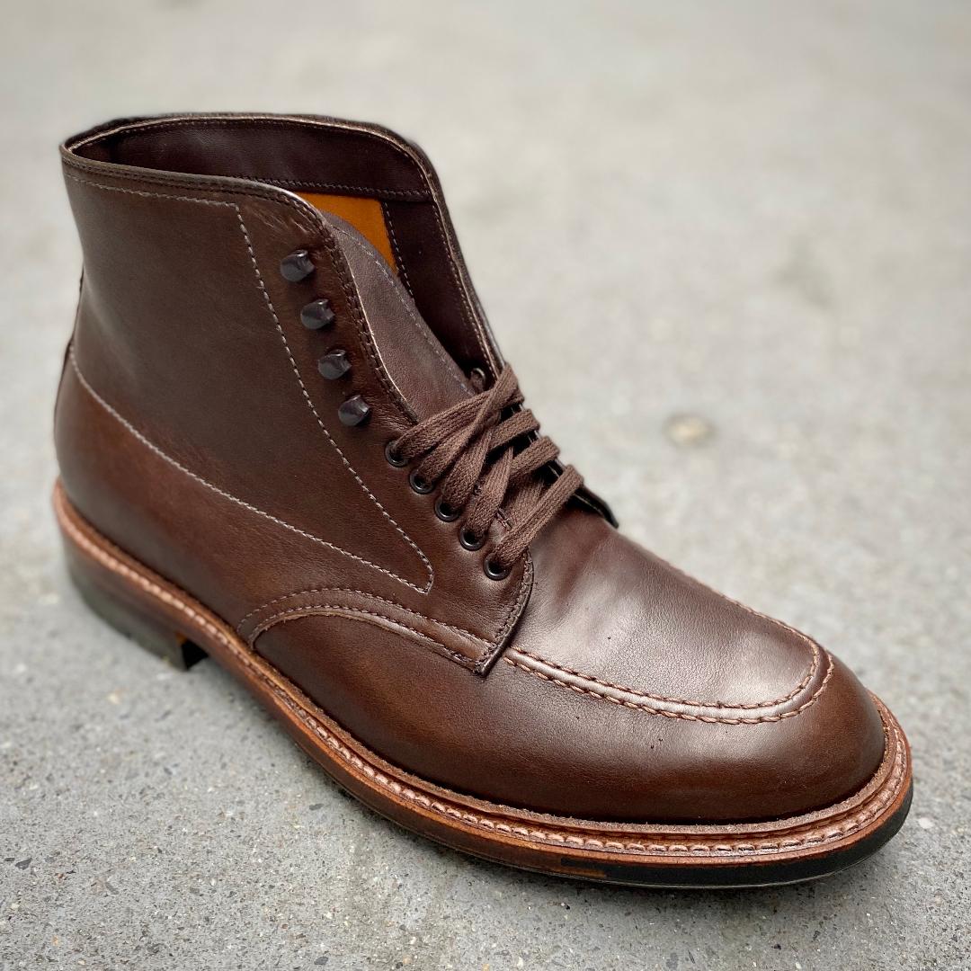 The Alden Madison 4012HC - Indy Boot in Brown Rusticalf, featuring dark brown laces, visible stitching, metal eyelets, a 360 Commando Sole, and a slightly raised heel, is displayed on a gray concrete surface.