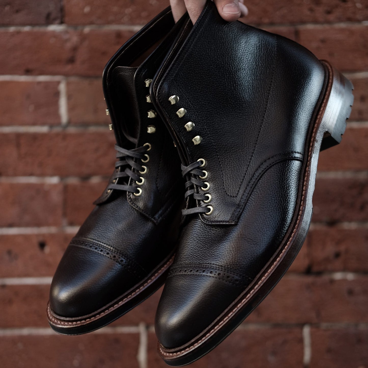 The D9819HC - Jumper Boot by Alden Madison is displayed against a textured red brick wall. These polished black leather boots, crafted from calfskin, feature brass eyelets, brown stitching, a cap toe design, dark laces, and a sturdy commando sole for added durability.