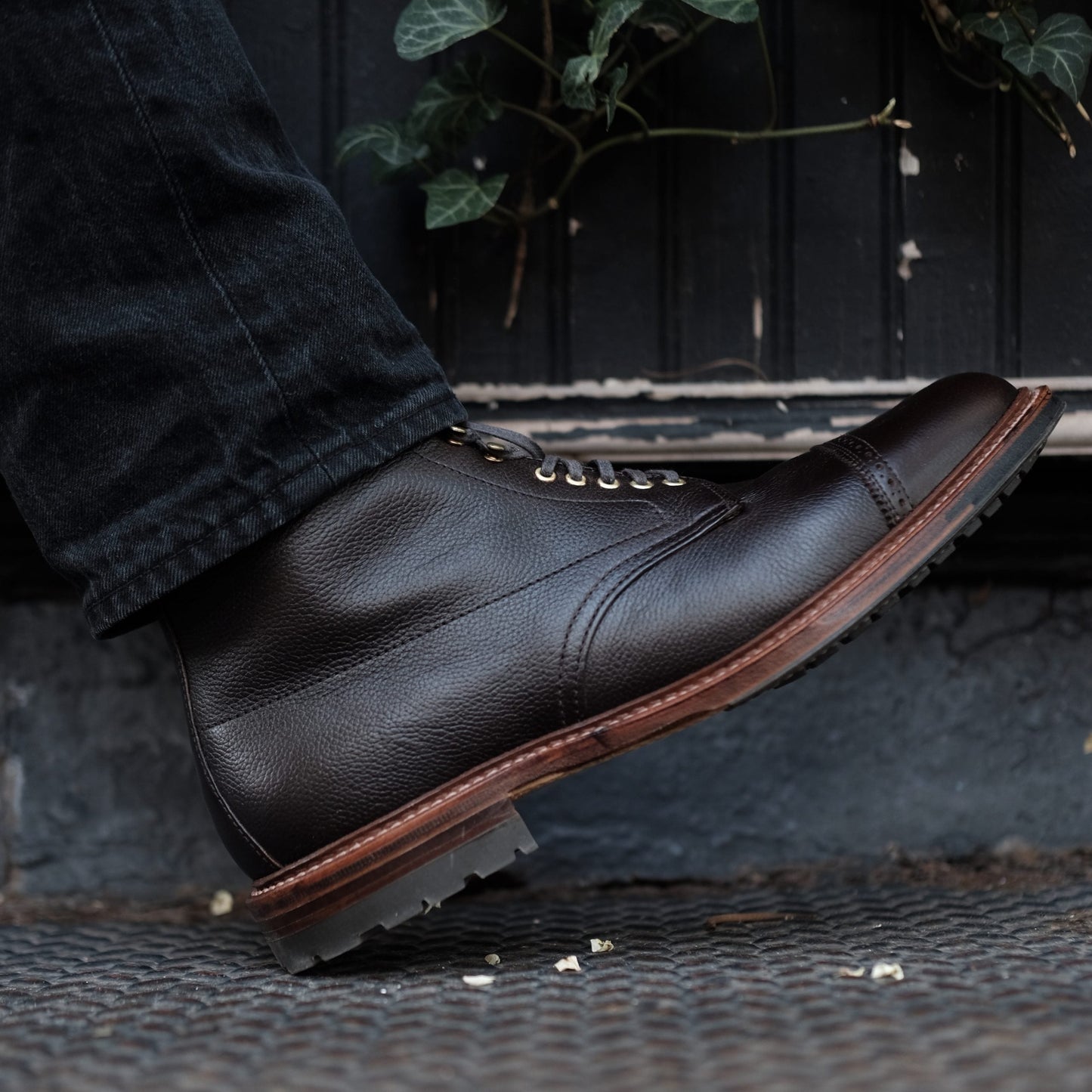 A close-up shows a person in Alden Madisons D9819HC - Jumper Boot in Regina Brown Grain standing on a patterned metal surface. The boots, crafted from premium calfskin, complement sleek dark pants above them. Lush green foliage peeks through in the background.