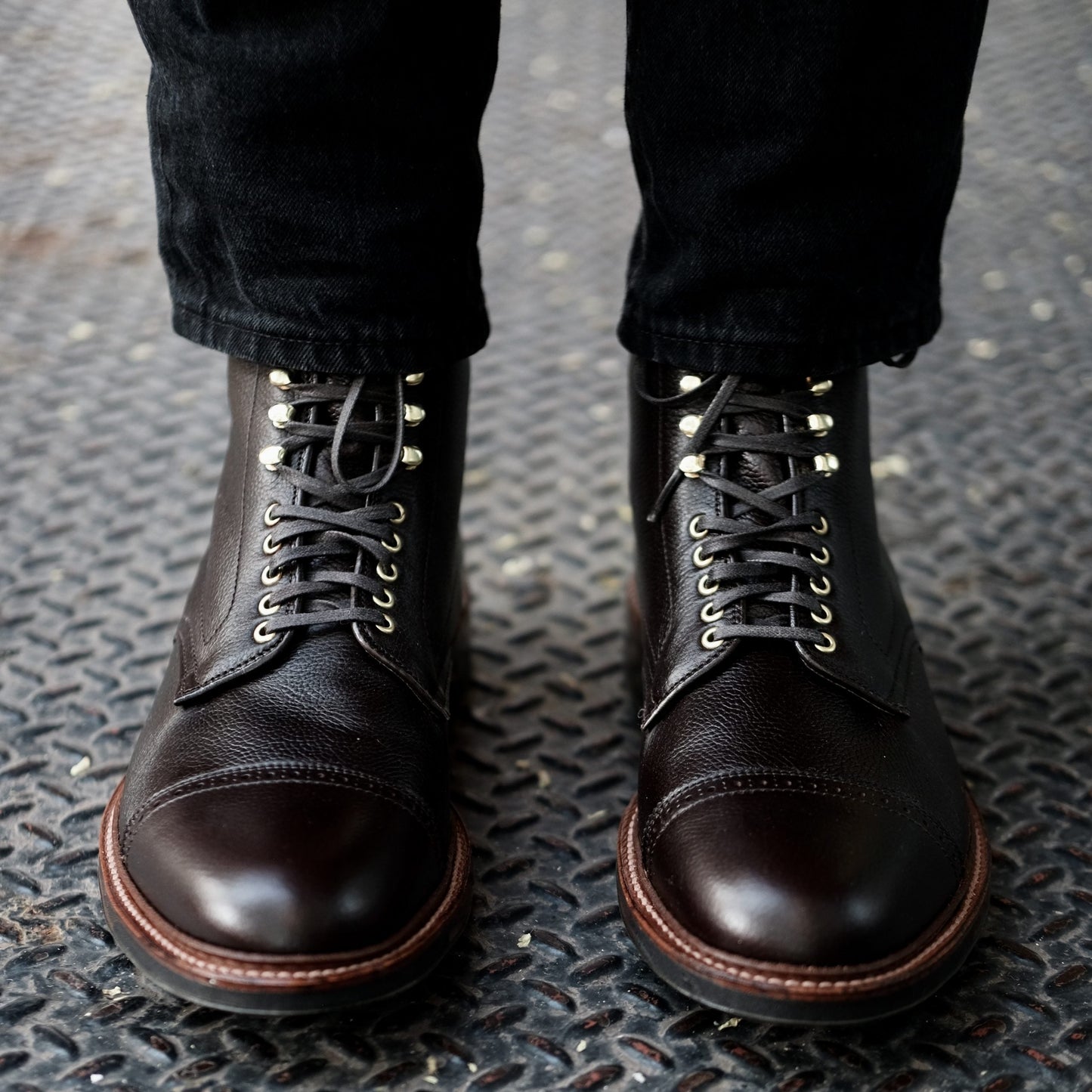 Dark brown Alden Madison D9819HC Jumper Boots in Regina Brown Grain calfskin leather, worn over black pants, feature metal eyelets, a smooth toe cap, and a rugged Commando Sole resting on a textured steel surface.
