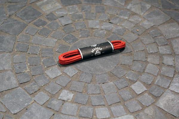 A coiled red Shoe Lace Guarded Goods, 27 Inch by Alden Madison, with a black and white label rests on a circular gray stone tile pattern.