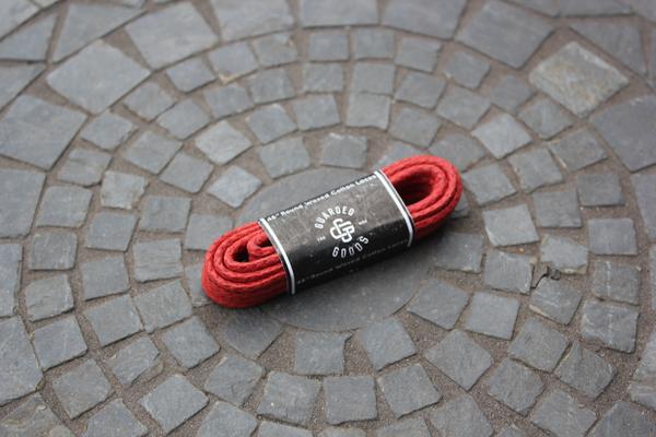 A coiled 48-inch Round Boot Lace by Alden Madison, thick and red with a black label featuring white text and logo, rests on circular cobblestone pavement. The spiral stone pattern around it imparts an air of guarded goods.