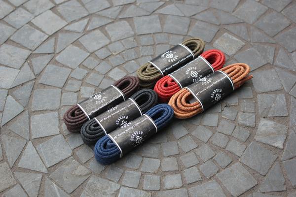 Six pairs of 48-inch Alden Madison Round Boot Laces in black, tan, olive, red, brown, and orange sit neatly coiled on a stone-tiled surface. Each pair is wrapped with a black band featuring the brand name.