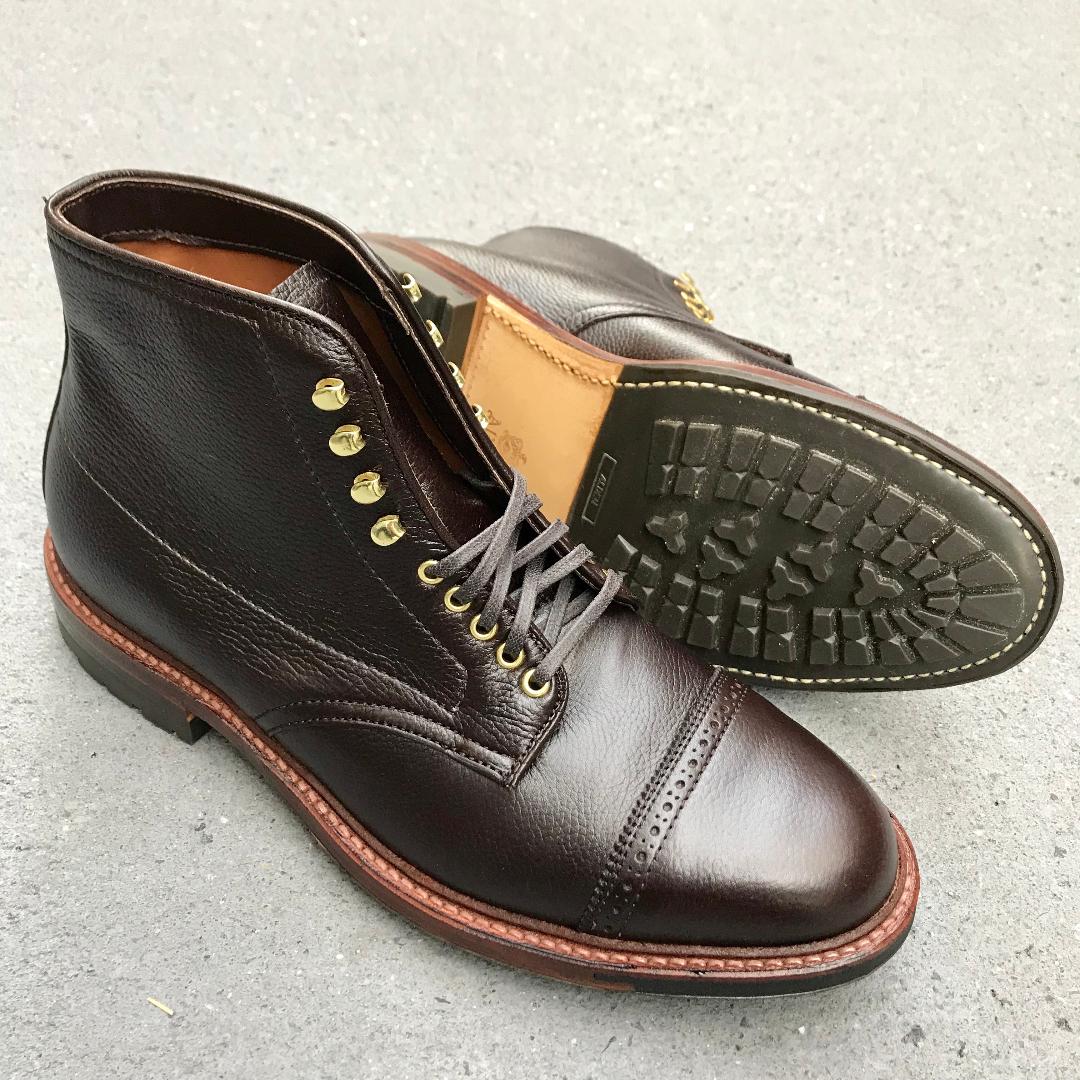 A pair of D9819HC - Jumper Boots in Regina Brown Grain by Alden Madison is displayed. One boot stands upright, the other lies on its side, highlighting the Commando Sole and contrasting red-brown welt stitch. Both rest on a gray surface with laces and gold eyelets.