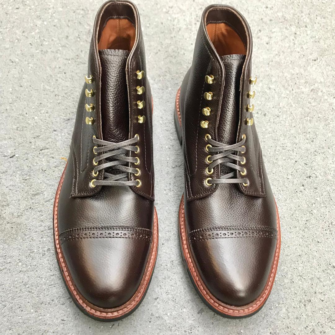 A pair of Alden Madisons D9819HC - Jumper Boot in Regina Brown Grain, featuring laces, decorative toe stitching, gold eyelets, a commando sole, and polished finish with brown soles, rests on a textured gray surface.