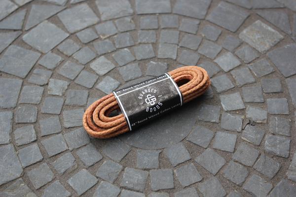 Round Boot Laces - 48 Inch by Alden Madison elegantly rest on a gray cobblestone surface with a circular pattern.
