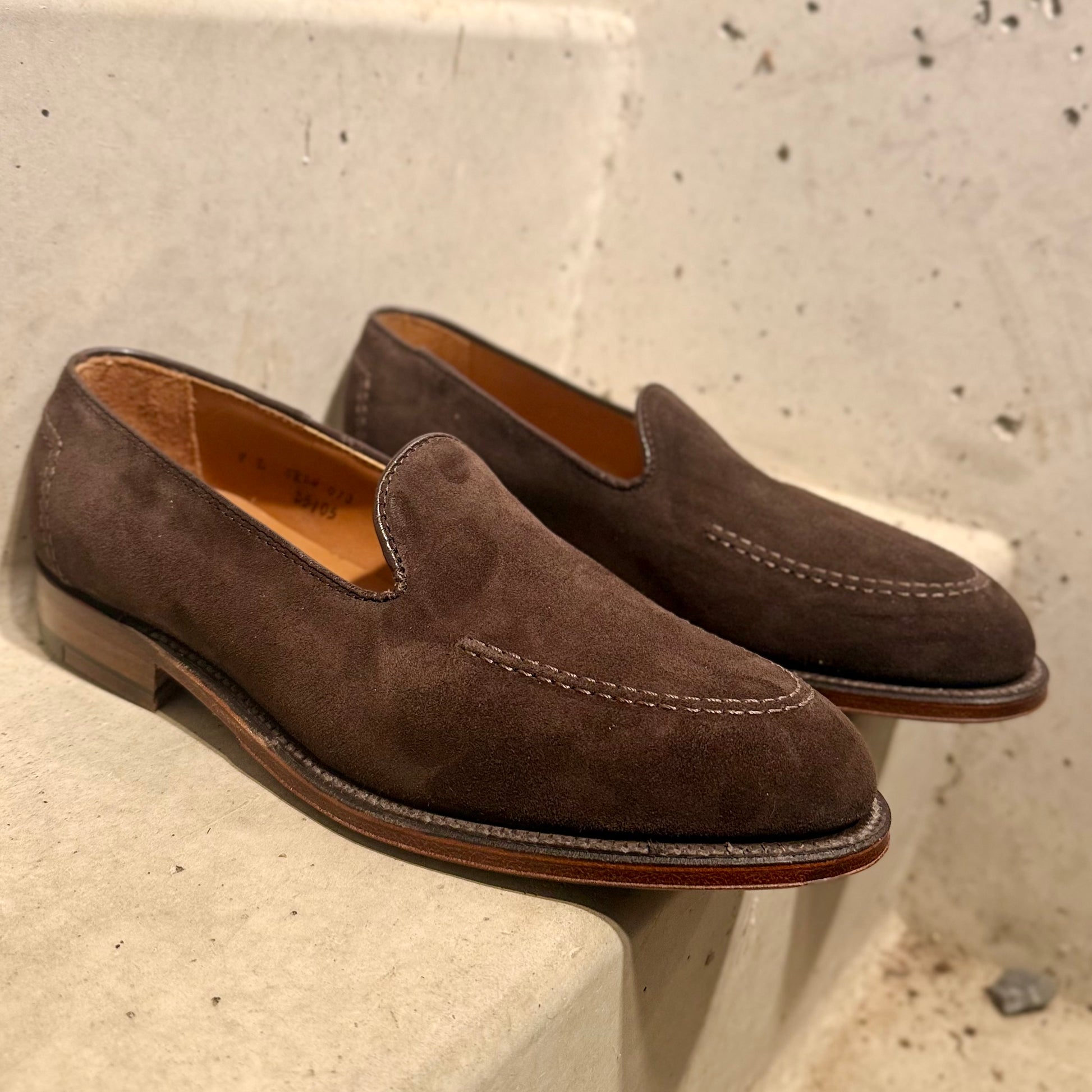 A pair of ALDEN SHOES D5105 - Vanderbilt in Dark Brown Suede, featuring visible stitching, sits on a light concrete step against a textured, speckled wall.