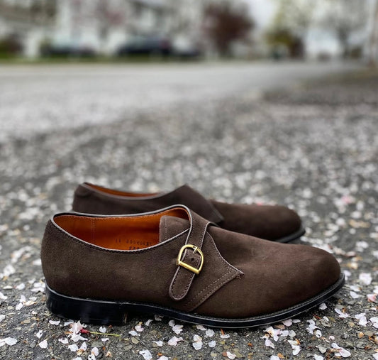 The Alden Madison 953 - Monk Strap in Brown Suede, with gold buckles, is displayed on outdoor pavement scattered with flower petals, set against a blurred background of trees and buildings.