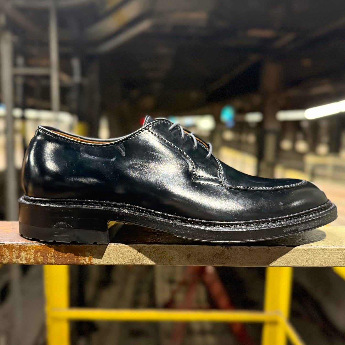 The ALDEN SHOES D4604C - Moc Toe “Borough” Blucher in Black Shell is shown on a wooden surface in an industrial space, with its visible stitching and lustrous finish set against blurred pipes and yellow railings.