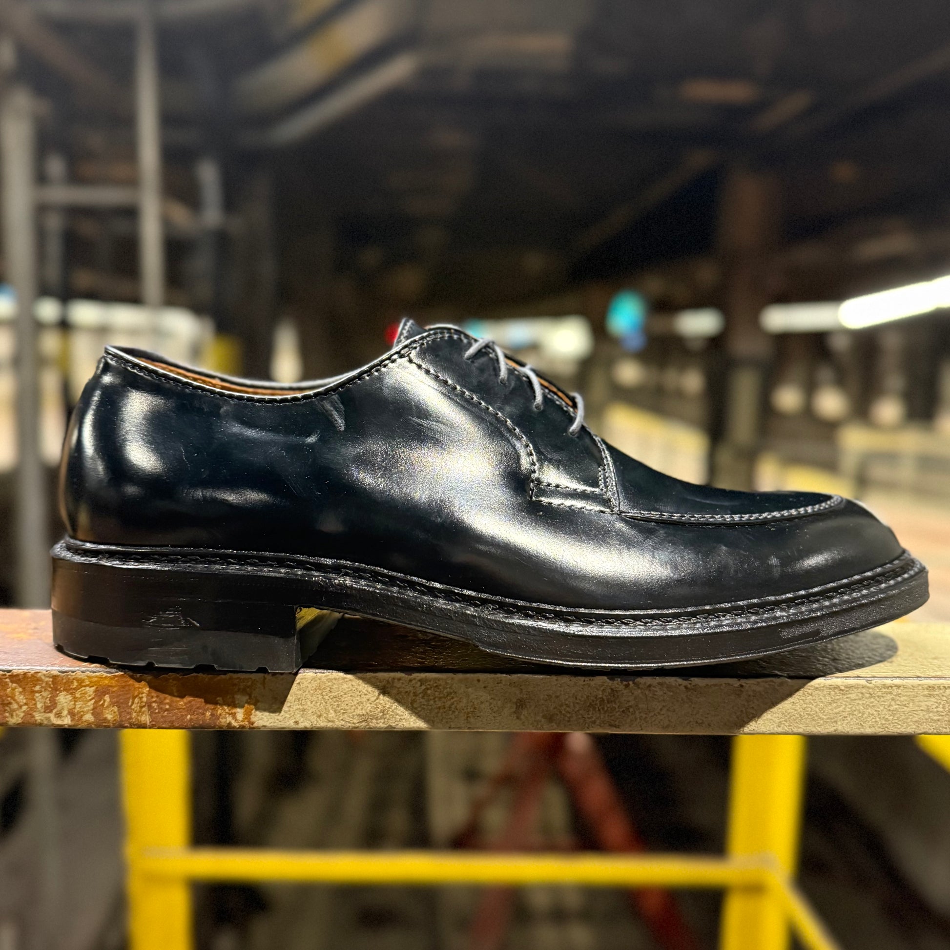 The ALDEN SHOES D4604C - Moc Toe “Borough” Blucher in Black Shell is shown on a wooden surface in an industrial space, with its visible stitching and lustrous finish set against blurred pipes and yellow railings.