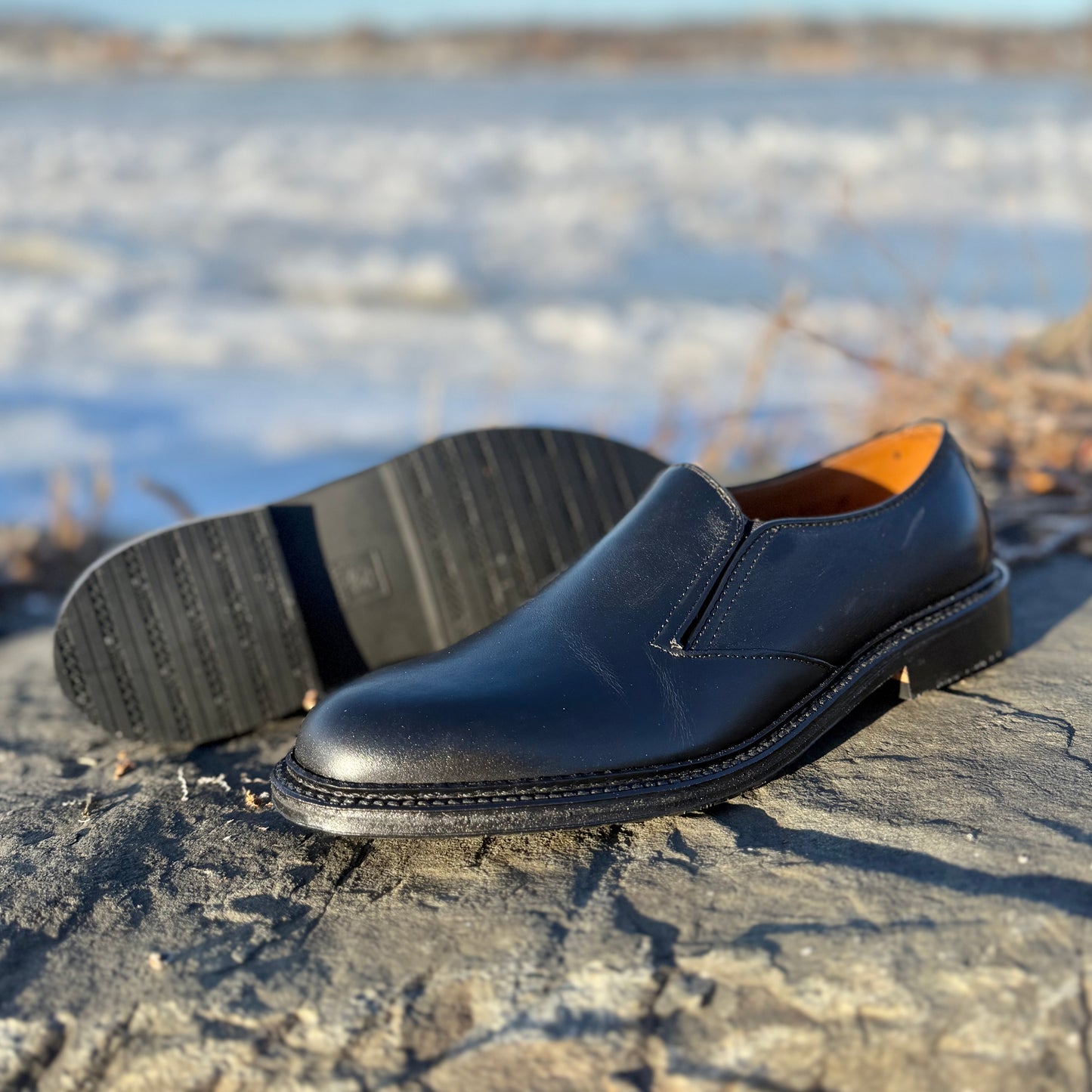 The ALDEN SHOES D2406L "Double Gore" black calfskin slip-on shoe is shown outdoors on a rock, with one upright and the other revealing its rubber sole, set against a blurred snowy background for an elegant effect.