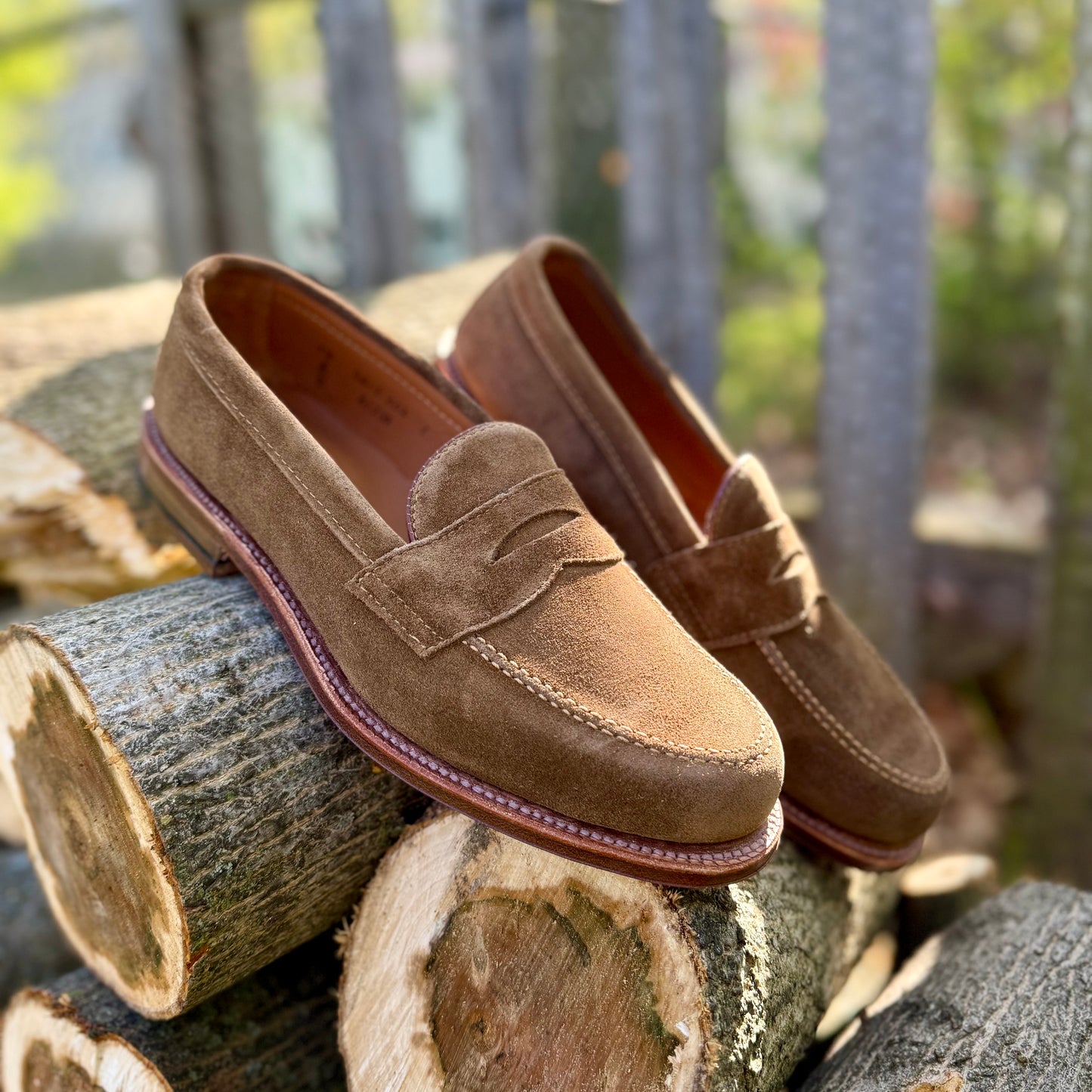 The Alden Madison 6173F - Penny Loafer in Snuff Suede, with stitched detailing and a Flex Leather Sole, sits outdoors on stacked tree logs as sunlight highlights its rich texture.