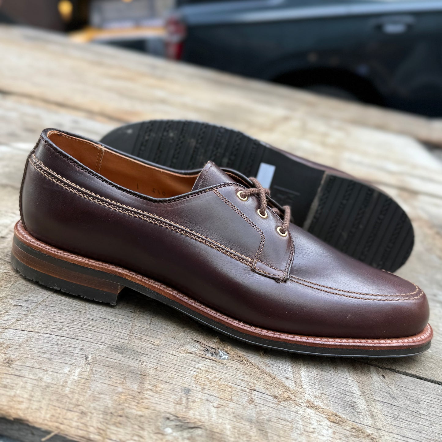 The Alden Madison 734L - Ivy Blucher in Brown Chromexcel, featuring visible stitching, laces, and a thick Leisure Rubber Sole, is shown on wood—one shoe flat, the other on the Van Last propped behind it.