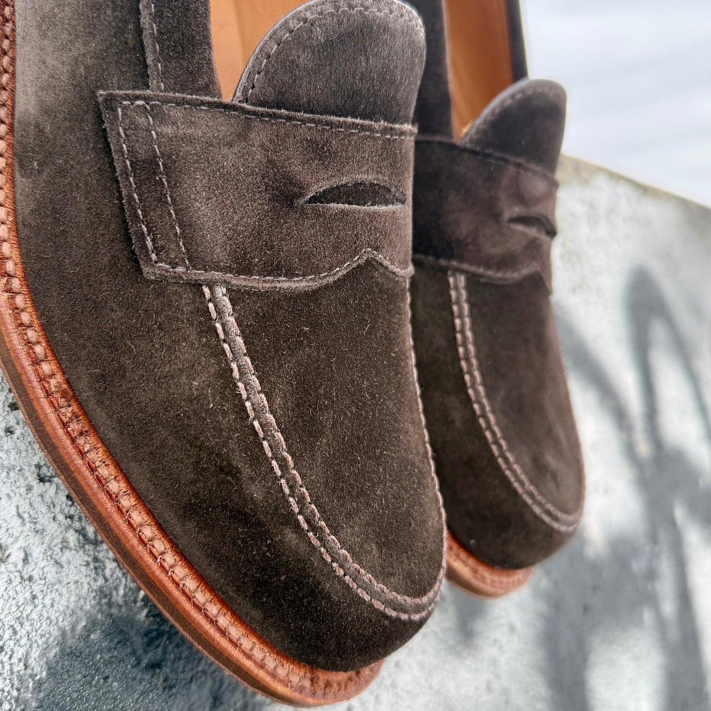 Close-up of Alden Madison's 6176F - Penny Loafer in Dark Chocolate Suede, featuring visible stitching and tan flex oiled leather soles, resting on a light gray textured surface.