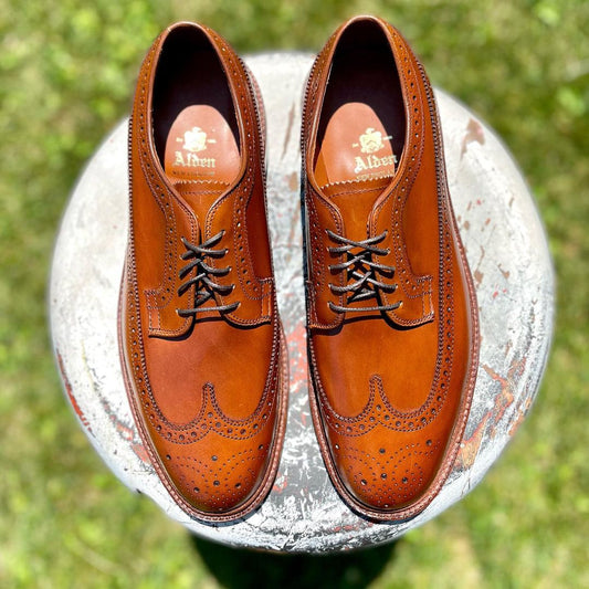 A pair of Alden Madison 979 Long Wing Bluchers in burnished tan calfskin, featuring intricate brogue detailing and a Double Oak Leather Outsole, rests on a weathered white stool outdoors. Dark laces complement the shoes, which showcase the label printed on the insole.