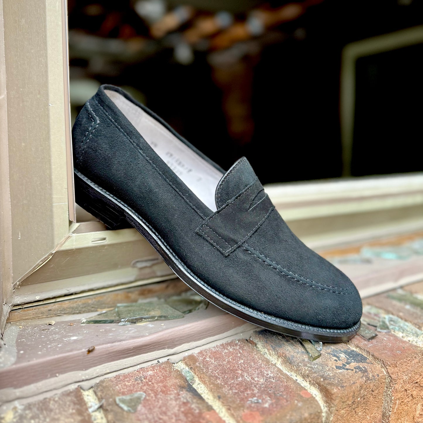 The Alden Madison D2229 “Stuyvesant” Madison Penny in Black Suede sits on a windowsill, angled to show its side and top view with a brick wall behind, emphasizing the loafer’s sleek leather sole.
