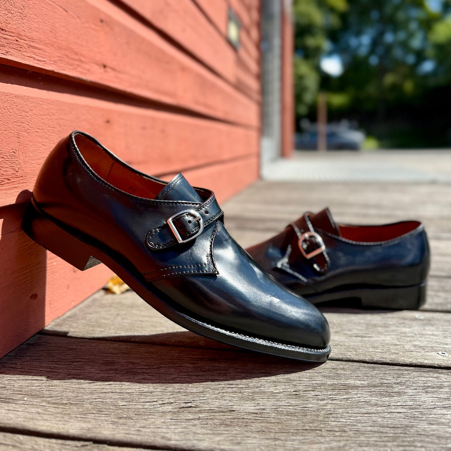 A pair of Alden Madison 1655 - Monk Strap Black in Shell Cordovan shoes with silver buckles rests on a wooden deck beside a red wall, green trees and sunlight softly blurred in the background.