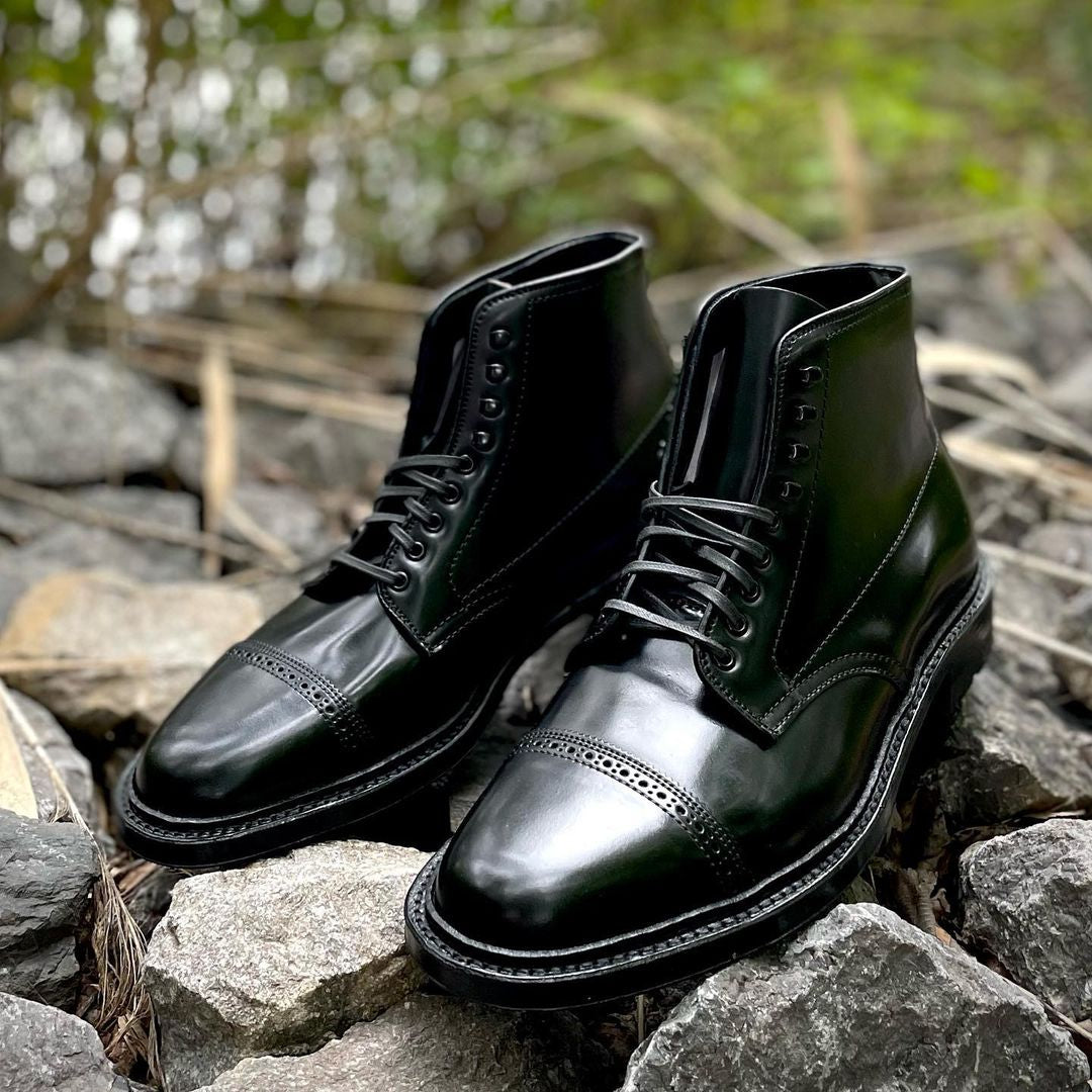 A pair of D7843C - Jumper Boots in Black Shell Cordovan from Alden Madison, featuring Speed Hook Lacing and detailed stitching, rests on gray rocks. With a sturdy Commando Sole, theyre perfect for outdoor adventures, as blurred greenery hints at the wild beyond.