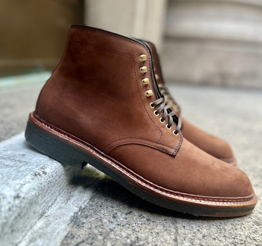 The Alden Madison 86032H Smooth Tobacco Chamois Plain Toe Boots, with crepe soles and eyelets, rest stylishly on a concrete surface. Featuring dark laces, their craftsmanship is highlighted by an elevated heel positioned on a curb.