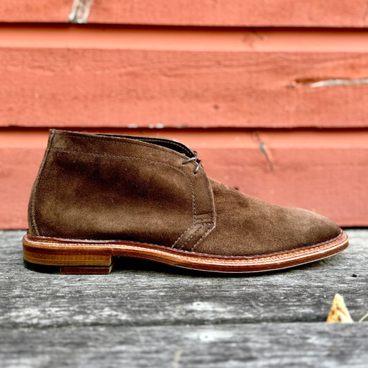 The Alden Madison 1492 - Unlined Chukka in Brown Suede features light brown stitching and a single flex leather sole, shown on a weathered wood surface against a red wooden wall.