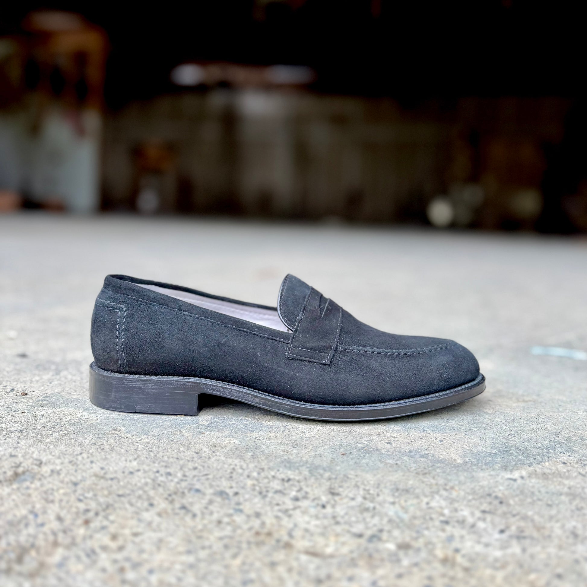 The Alden Madison D2229 “Stuyvesant” Madison Penny in black suede, with a leather sole, is displayed in profile on concrete, facing left against a blurred dark background.