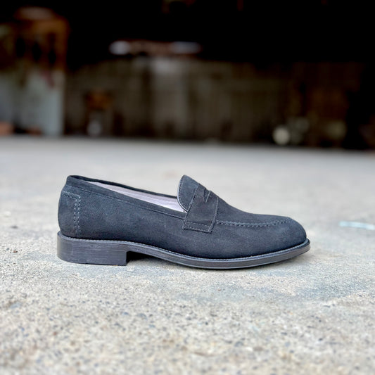 The Alden Madison D2229 “Stuyvesant” Madison Penny in black suede, with a leather sole, is displayed in profile on concrete, facing left against a blurred dark background.
