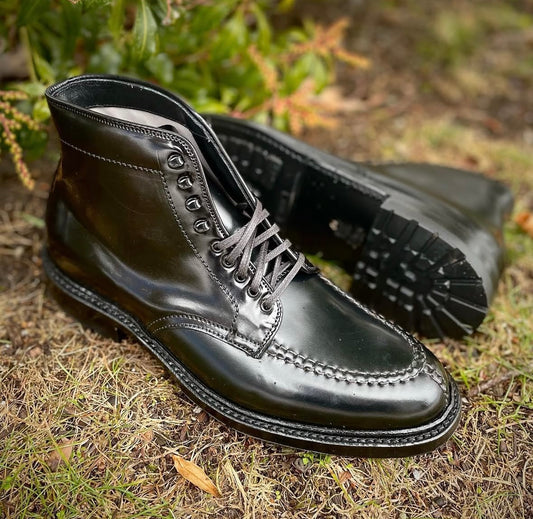 Alden Madisons 40509HC Indy Boot w Commando Black Shell Cordovan, a pair of shiny black lace-up boots with rugged Commando soles, shown outdoors on grass and dirt with green foliage in the background.