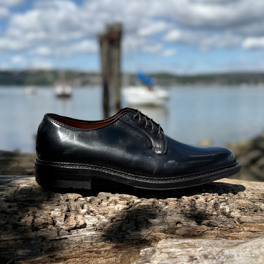 The Alden Madison 9901C Plain Toe Blucher in Black Shell Cordovan with a Commando Sole rests on a weathered wooden log, set against a blurred backdrop of water, boats, and a cloudy sky.
