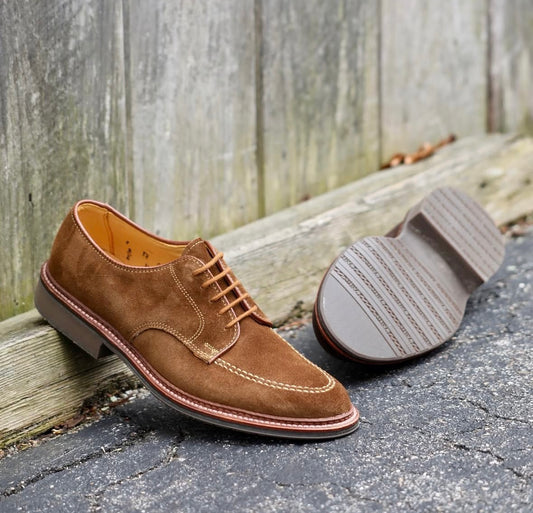 Alden Madison’s 702 - Mocc Toe Blucher in Snuff Suede, featuring a lace-up design and Dress Rubber Sole on the Orleans Last, is displayed outdoors—one upright and one showing the sole—on a paved surface beside a wooden fence.
