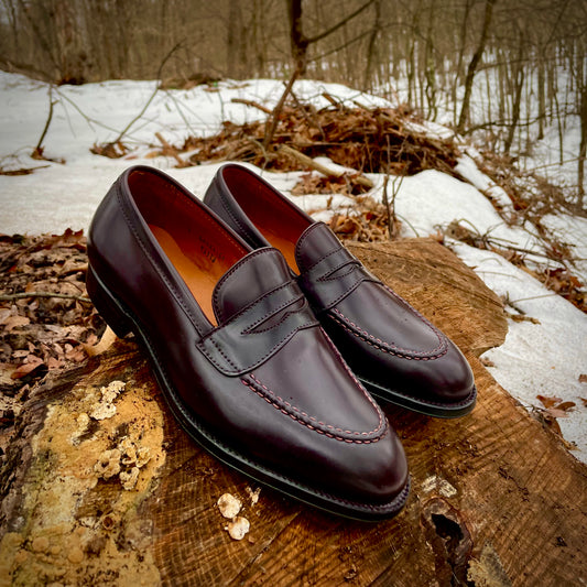 A pair of Alden Madisons 67169 - High Vamp Loafers in Color 8 Shell Cordovan leather, crafted on the Grant Last, is artfully displayed on a tree stump in a snowy forest. The polished shoes feature exquisite hand stitching amid scattered leaves and snow-covered ground with bare trees.