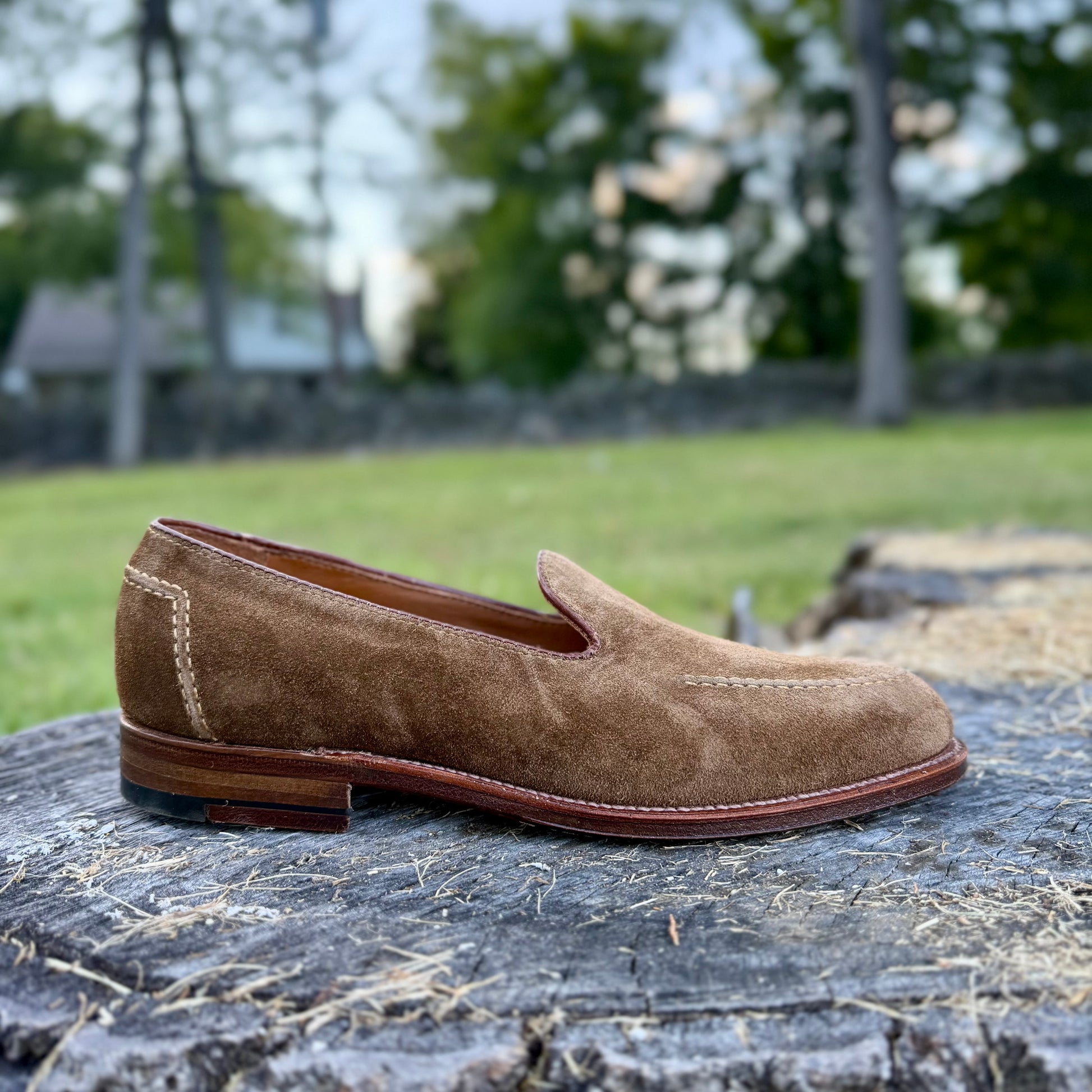 The Alden Madison D2101 - Vanderbilt Slip On in snuff suede, featuring visible stitching and a leather sole, rests on a tree stump outdoors with blurred grass, trees, and a stone wall in the background.
