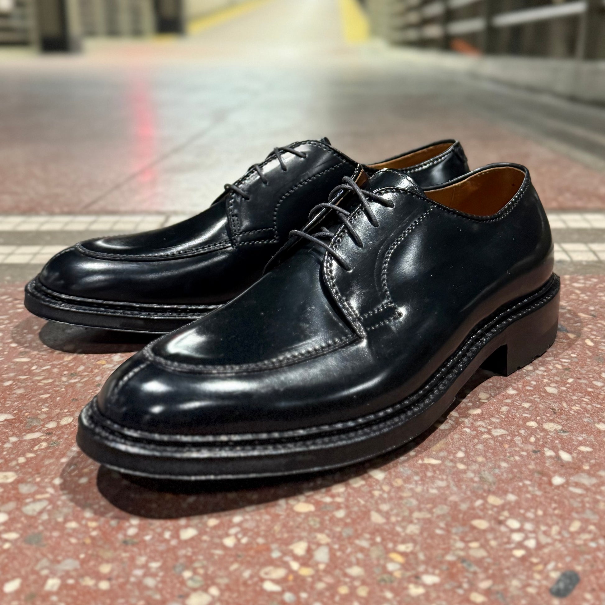 The ALDEN SHOES D4604C - Moc Toe “Borough” Blucher in Black Shell features thick soles and visible stitching in a classic style, shown on a speckled tile floor under bright indoor lighting.