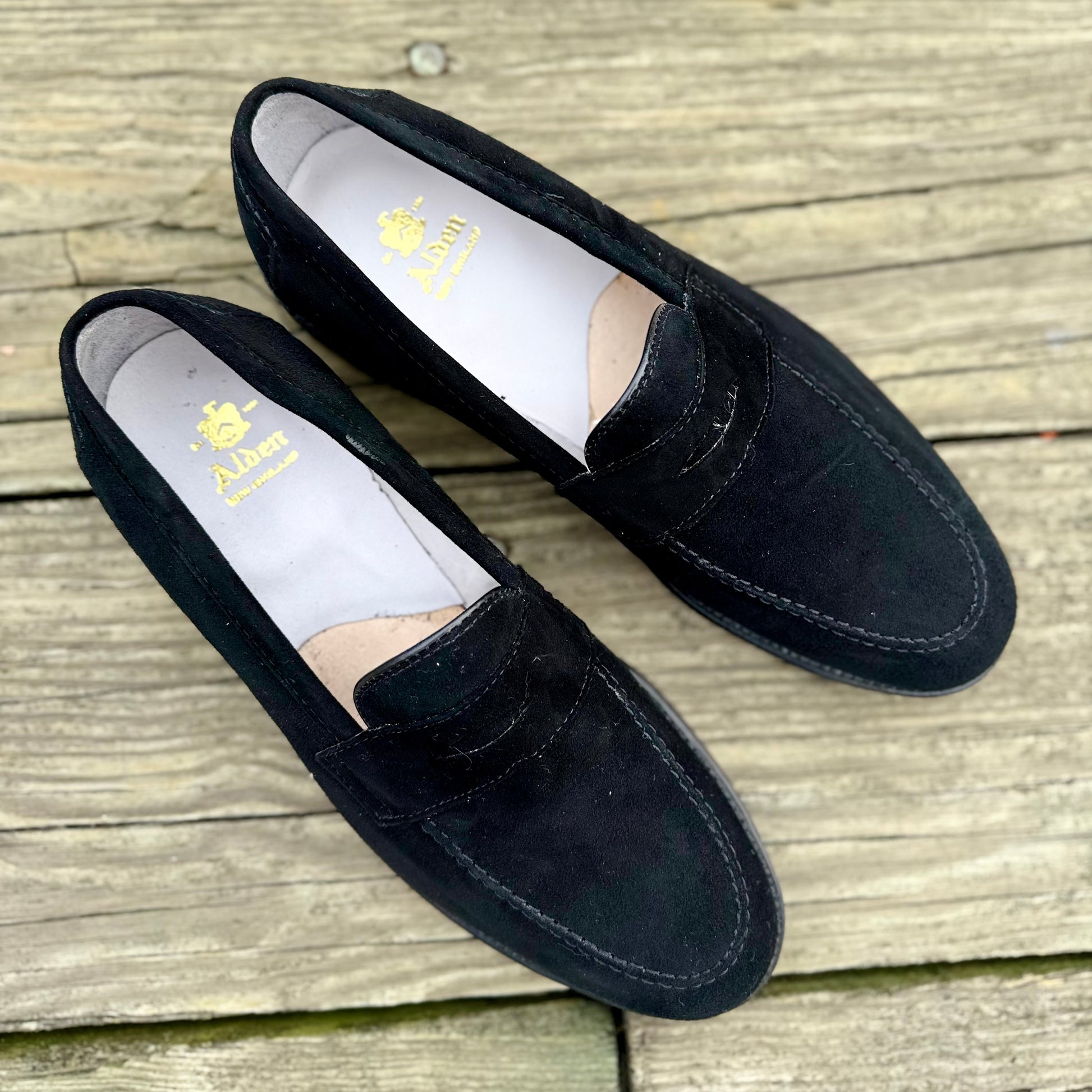 A pair of Alden Madison D2229 “Stuyvesant” Madison Penny loafers in black suede, featuring gold insole branding and leather soles, displayed on weathered wooden planks.