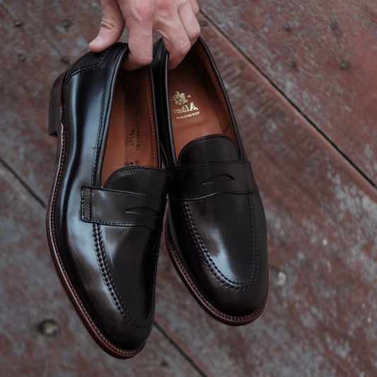 A hand holds a pair of shiny dark brown leather Alden Madison Penny loafers (96288) in Color 8 Shell Cordovan against a rustic wooden background, showcasing the stitching details, rounded toes, antique edging, and visible brand logo on the insole.