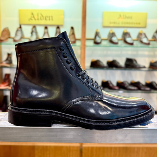 The Alden Madison D6842H - Plain Toe Director Boot in Black Shell Cordovan, crafted on a Barrie Last, is laced and displayed on a wooden surface in a shoe store, with shelves of various shoes in the background.