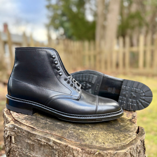 The Alden Madison 40797HC Black Cap Toe Boot with Commando Sole is shown outdoors on a tree stump—one boot upright, the other on its side to display the sole—against a softly blurred background of trees and a wooden fence.