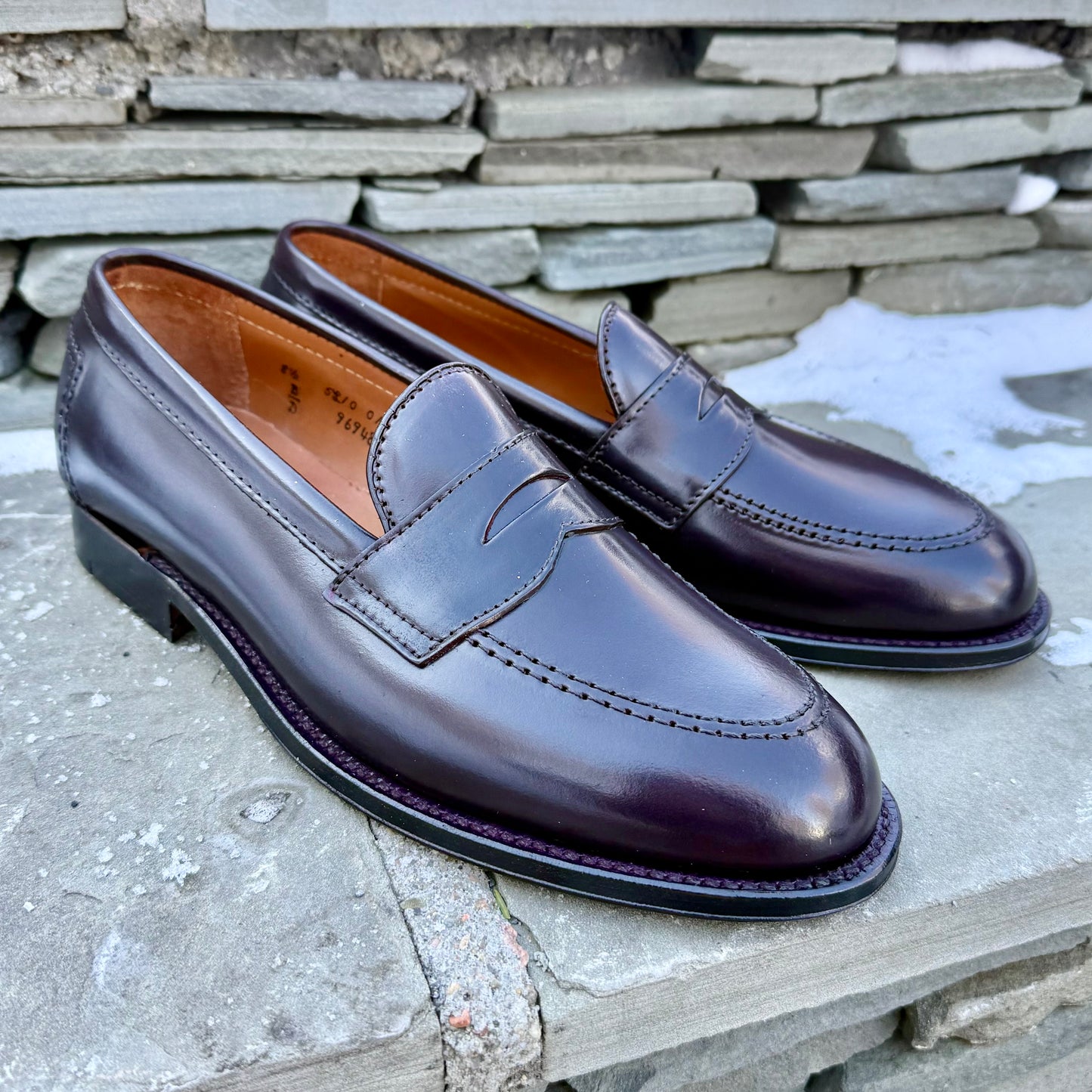 A pair of ALDEN SHOES 96948 - Copley Penny Loafer in Color 8 Shell sits on a stone surface, framed by stacked stone and patches of snow.