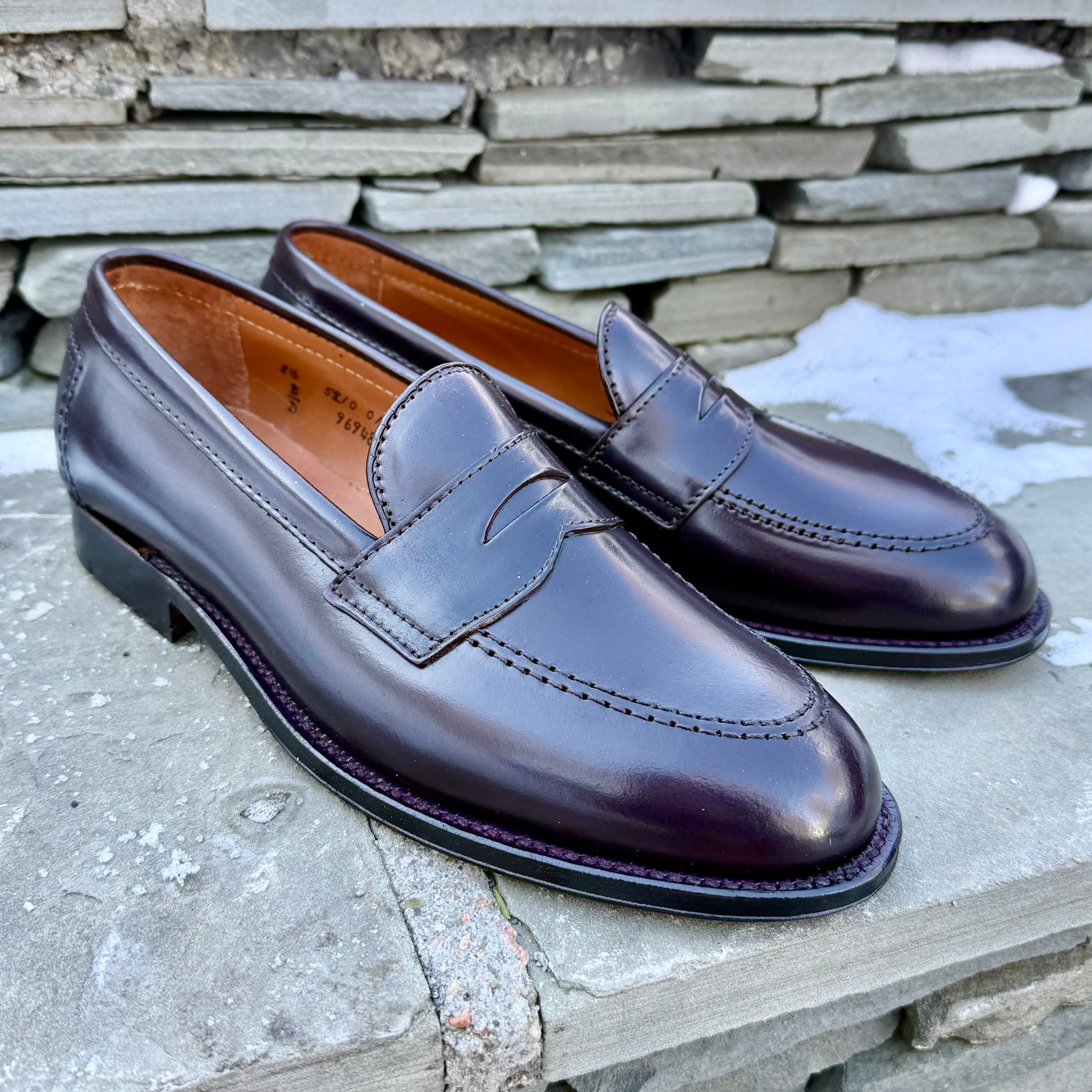 A pair of ALDEN SHOES 96948 - Copley Penny Loafer in Color 8 Shell sits on a stone surface, framed by stacked stone and patches of snow.