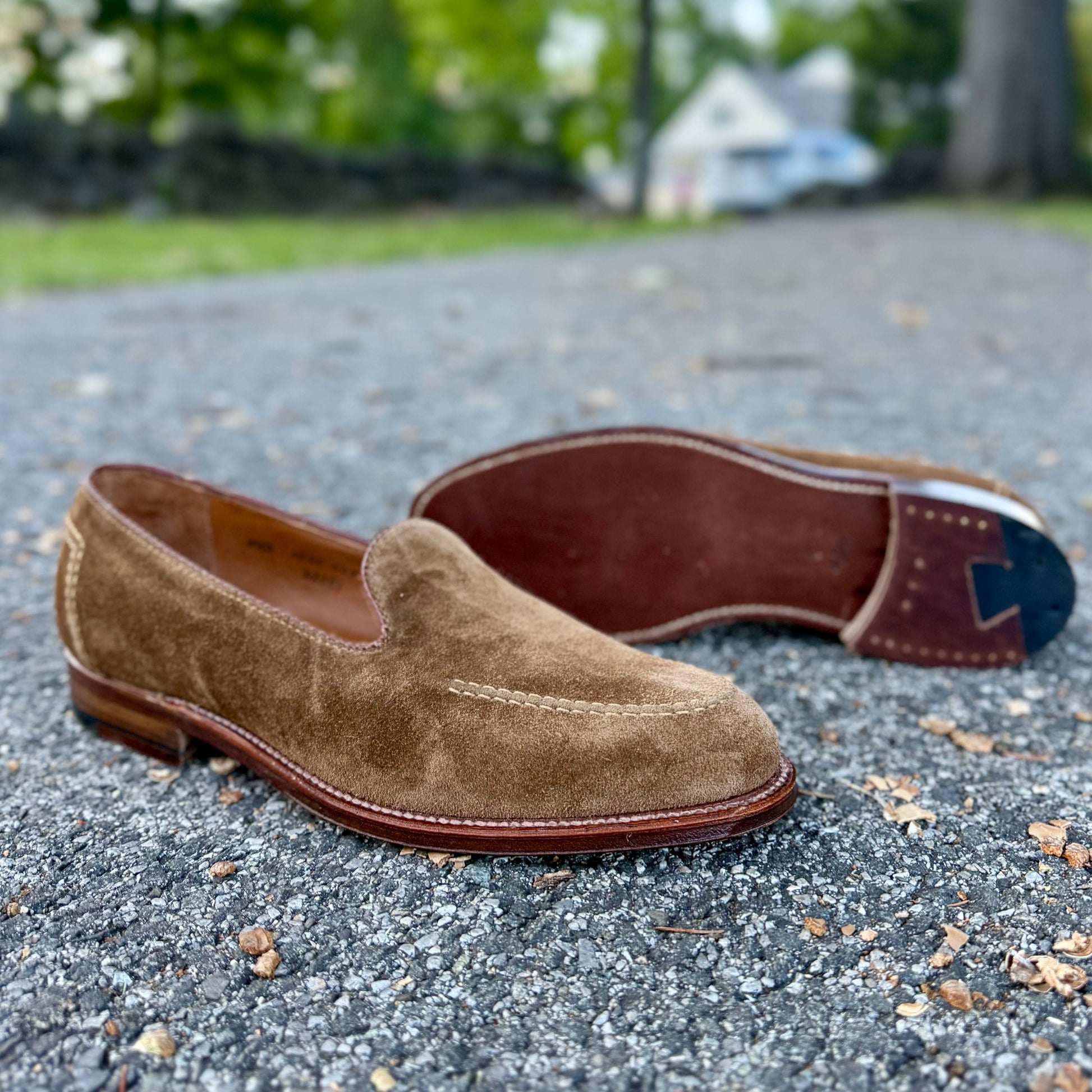 A pair of Alden Madison D2101 - Vanderbilt Slip On in snuff suede sit on the pavement, crafted with sturdy leather soles; one shoe stands upright, the other lies on its side with greenery and a house blurred in the background.