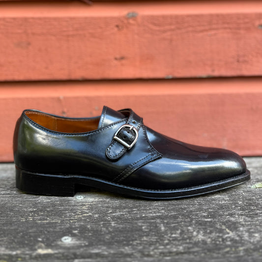 The Alden Madison 1655 - Monk Strap Black in Shell Cordovan dress shoe, featuring a silver buckle, rests on a weathered wooden surface with a red wooden wall in the background.