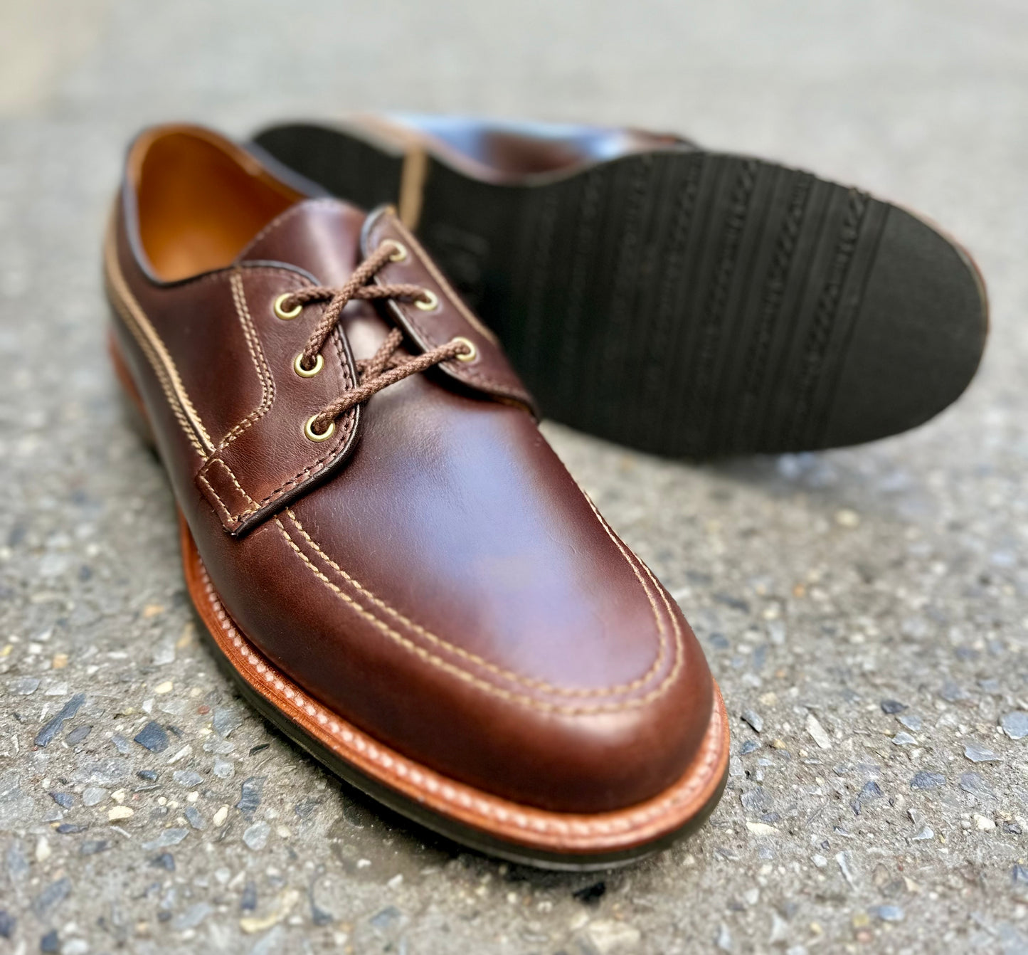 A close-up of the Alden Madison 734L - Ivy Blucher in Brown Chromexcel shows detailed stitching, one shoe upright and the other revealing its black leisure rubber sole, displayed on a textured gray surface.