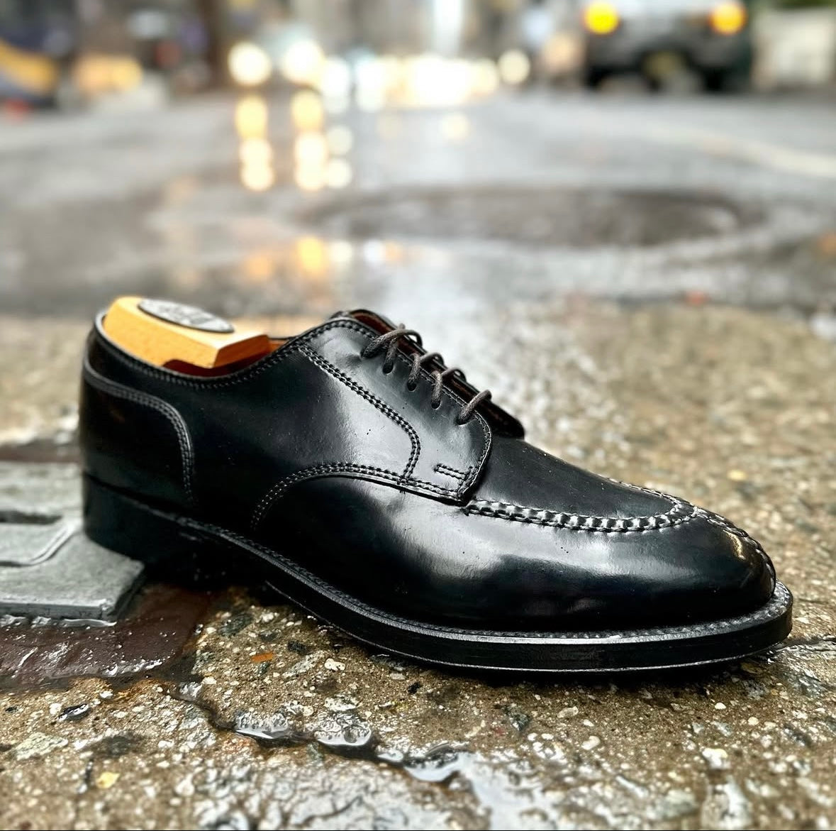 A close-up of the Alden Madison 2211 Norwegian Split Toe Blucher in Black Shell Cordovan shows black laces and a double oak leather outsole, set on wet city pavement with blurred lights and cars in the background.