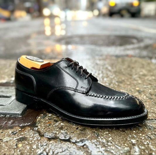 A close-up of the Alden Madison 2211 Norwegian Split Toe Blucher in Black Shell Cordovan shows black laces and a double oak leather outsole, set on wet city pavement with blurred lights and cars in the background.