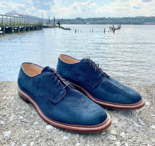 The Alden Madison 29331F Unlined Plain Toe Blucher Blue Suede shoes are side by side on concrete near water, with a pier and shoreline in the distance under a partly cloudy sky.