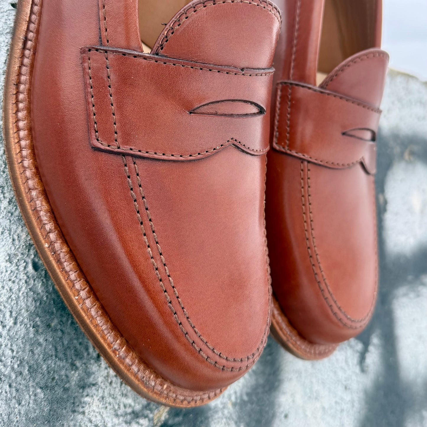 Close-up of the Alden Madison 9823 Penny Loafer in Tan Michigan Calf, featuring visible stitching and an Oak Leather Sole, shown polished on a textured gray surface.